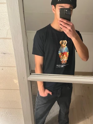 Mörkblå Polo Bear t-shirt från Ralph Lauren - Mörkblå t-shirt från Polo Ralph Lauren med ikoniskt Polo Bear-tryck på bröstet. Klassisk passform och rund halsringning. Tillverkad i mjuk bomull, perfekt för en avslappnad och stilren look, passar också S.