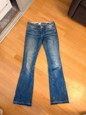 Jeans låg midja Mavi  - Säljer ett par blå bootcut jeans med slitningar och råa kanter nertill. Jeansen har klassisk femficksdesign, låg midja och ljusa tvättade partier framtill och baktill. Perfekta för en avslappnad och trendig look.