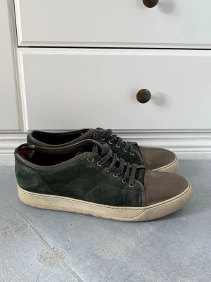 Mörkgröna Lanvins - Snygga sneakers i mörkgrön mocka med bruna läderdetaljer på tå och snörning. Skorna har en vit sula och klassisk låg profil. Perfekta för dig som gillar stilrena och tidlösa sneakers med en twist. Defekt på hälen.