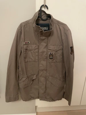 Beige jacka från Herno med patchar - Snygg beige jacka från Herno med coola patchar på bröstet och ärmen. Jackan har hög krage, fyra fickor framtill och stängs med dragkedja och knappar. Perfekt för dig som gillar en stilren och avslappnad look.