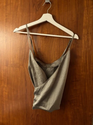 Grå omlott linnetopp med smala band - Snygg grå omlott linnetopp i satin med smala axelband. Toppen har en draperad v-ringning och är luftig med en stilren look. Perfekt för att lyfta vilken outfit som helst.