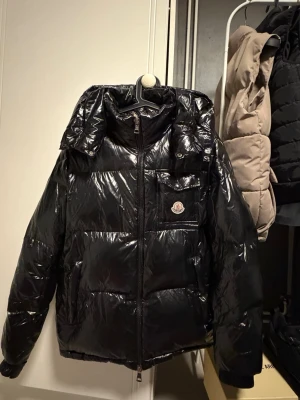 Svart glansig dunjacka från Moncler - Snygg svart dunjacka från Moncler med glansigt yttertyg, huva och dragkedja framtill. Jackan har en bröstficka med lock och Moncler-logga samt vita och röda detaljer längs insidan av dragkedjan. Perfekt för kalla vinterdagar.