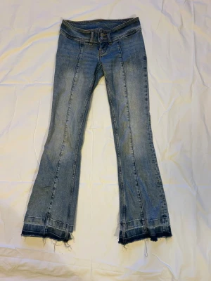 Blå bootcut jeans med råa fållar - Säljer ett par ljusblå bootcut jeans med markerade sömmar framtill och råa, fransiga fållar längst ner. Jeansen har låg midja och klassisk femficksdesign. Materialet är denim i bomull. Perfekta för en avslappnad och trendig look.