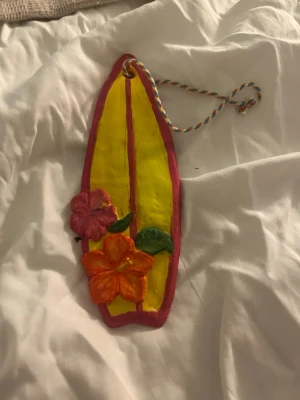 Dekorativ surfbräda i keramik - Färgglad hängande dekoration formad som en surfbräda i keramik, målad i gult och rosa med två blommor i orange och rosa samt gröna blad. Har ett snöre för upphängning. Perfekt för att ge rummet en tropisk vibe.