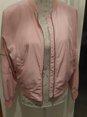 Ljusrosa bomberjacka med dragkedja - Snygg ljusrosa bomberjacka med klassisk passform och ribbade muddar. Jackan har dragkedja framtill, fickor på sidorna och en ärmficka med dragkedja. Tillverkad i ett glansigt material som ger en cool look. Tyvärr 3 tunna vita korta streck på ena armen.