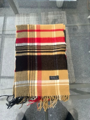 Burberry halsduk – klassisk rutig design i 100% lambswool - Säljer en äkta Burberry halsduk i det ikoniska klassiska rutmönstret. En tidlös designerhalsduk tillverkad i mjuk och varm 100% lambswool (ull) – perfekt för höst, vinter och kalla vårdagar.  Den klassiska rutiga designen i beige, camel, svart, rött och vitt gör den lätt att matcha med trenchcoat, ullkappa, blazer eller vardagsjacka. En stilren lyxaccessoar som funkar lika bra till preppy outfits, old money-stil som modern streetwear. Passar både dam och herr – unisex.  Detta är en äkta premium 