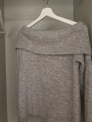 Ljusgrå off shoulder stickad tröja - Mysig ljusgrå stickad tröja med bred off shoulder-krage och ribbstickade detaljer. Perfekt för lager-på-lager och chill dagar. Tröjan har en avslappnad passform och är tillverkad i ett mjukt, melerat garn.