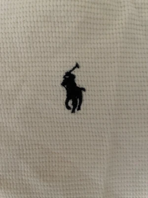 Vit t-shirt från Polo Ralph Lauren - Klassisk vit t-shirt från Polo Ralph Lauren med det ikoniska broderade polospelar-loggan på bröstet. T-shirten är kortärmad och har en ribbad struktur som ger en snygg och unik känsla. Perfekt till jeans eller shorts för en clean och stilren look.