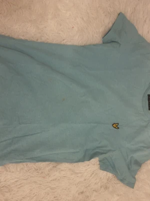 Ljusblå t-shirt från Lyle & Scott - Ljusblå t-shirt från Lyle & Scott med klassisk passform och korta ärmar. T-shirten har en gul broderad logga på bröstet och är tillverkad i mjukt bomullsmaterial. Perfekt för en avslappnad stil.