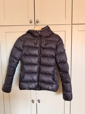 Svart dunjacka från Moncler - Snygg svart dunjacka från Moncler med huva och dragkedja framtill. Jackan har Moncler-logga på ärmen och diskreta detaljer på kragen. Puffig modell som håller värmen och passar perfekt för kalla vinterdagar.