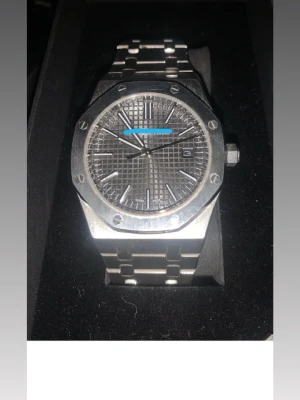 Audemars Piguet Royal Oak klocka - Säljer en Audemars Piguet Royal Oak klocka i silverfärgat stål med ikonisk åttakantig boett och länkarmband. Urtavlan är grå med rutmönster och har silverindex samt datumvisning. Snygg och tidlös design med automatiskt urverk som syns genom baksidan.