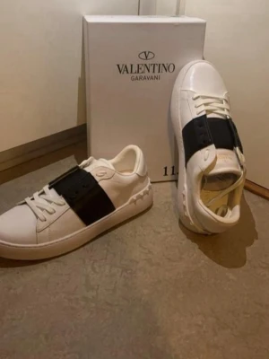 Valentino Garavani vita sneakers - Snygga vita sneakers från Valentino Garavani med en bred svart läderdetalj över vristen. Klassisk låg modell med vita snören och diskret logga på plösen och innersulan. Skorna har en stilren och modern design i lyxigt läder.