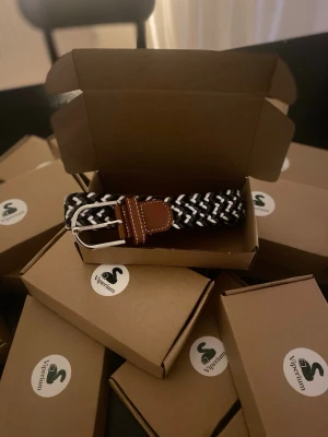 Svartvitt flätat bälte med läderdetalj - Snyggt svartvitt flätat bälte från Vipremium med silverfärgat spänne och bruna läderdetaljer vid spännet. Bältet har en elastisk design som gör det bekvämt och flexibelt att bära. Perfekt accessoar för att lyfta din outfit.
