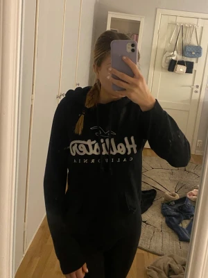 Svart Hollister hoodie med tryck - Säljer en svart hoodie från Hollister med vitt tryck på bröstet och klassisk logga. Hoodien har huva med dragsko och en stor magficka framtill. Perfekt för chill dagar. Materialet är mjukt och bekvämt, passar dig som gillar enkel och snygg design.