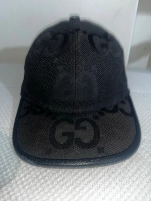 Svart keps från Gucci med GG-mönster - Snygg svart keps från Gucci med klassiskt GG-monogram över hela kepsen. Kepsen har böjd skärm och detaljer i svart läder runt kanten. Tillverkad i canvasmaterial med justerbar rem baktill. Perfekt för dig som gillar exklusiva accessoarer och streetstyle. De är borta typ den där bak den man fixar längden med och de står gucci på den där ute 