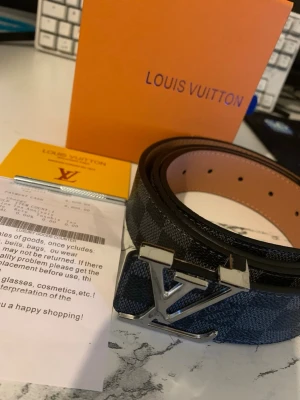 L,V rutigt bälte i skinn - Snyggt L och V bälte i svart och grått med rutigt mönster och ikoniskt L,V-spänne i silver. Bältet är gjort i skinn och har exklusiv känsla, perfekt för att lyfta vilken outfit som helst.