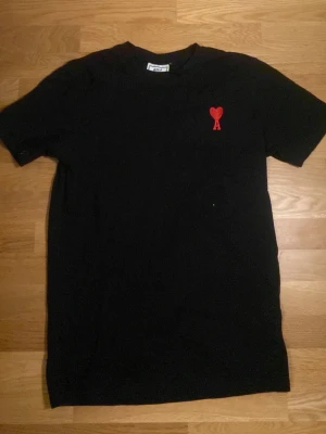 Svart t-shirt från Ami Paris med rött hjärta - Svart t-shirt från Ami Paris med klassisk passform och korta ärmar. På bröstet sitter Ami:s ikoniska röda hjärta med bokstaven A broderat. Tillverkad i mjuk bomull som känns skön mot huden. Perfekt till jeans eller shorts. Passar även S