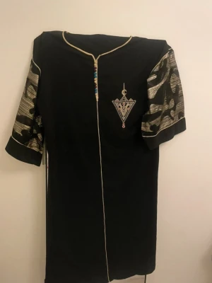 Svart klänning med broderi och mönstrade ärmar - Svart klänning med guldfärgade detaljer och broderad trekant på bröstet. Ärmarna är korta och har ett unikt mönster i guld och svart. Klänningen har en dekorativ kant längs halsringningen och framtill.