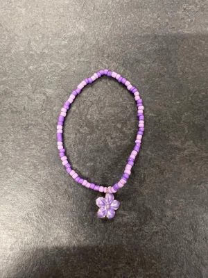 Lila pärlarmband med blomberlock storlek L - Ett sött armband med lila pärlor och ett hängande blomformat berlock. 