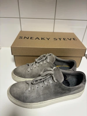 Gråa sneakers i mocka från Sneaky Steve - Snygga grå sneakers från Sneaky Steve i mjuk mocka med klassisk låg profil och vita sulor. Skorna har matchande grå skosnören och en stilren design som passar till det mesta. Perfekta för dig som gillar en clean och enkel look.