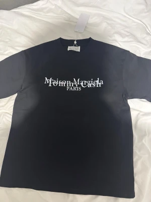 Maison Margiela svart t-shirt - Svart t-shirt från Maison Margiela med vit, stilren logotyp och text på bröstet samt ikonisk siffergrafik på ryggen. Rak passform och rund halsringning. Perfekt för dig som gillar exklusiva streetwear-plagg.