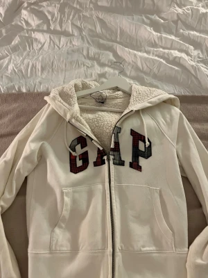 Vit hoodie med dragkedja från GAP - Mysig vit hoodie från GAP med dragkedja och fodrad insida i teddy. Stor GAP-logga framtill i röd- och svartmönstrad stil. Utrustad med huva och två fickor framtill. Perfekt för en chill och avslappnad look.