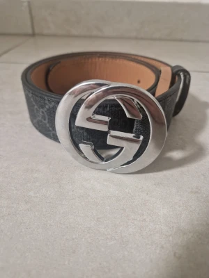 Gucci svart bälte med silverdetalj - Snyggt svart bälte från Gucci med klassiskt GG-mönster och stor rund silverspänne. Bältet har en lyxig känsla med detaljer i skinn och passar perfekt för att lyfta vilken outfit som helst.