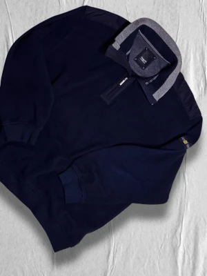 Mörkblå ziptröja från Bugatti - Mörkblå tröja från Bugatti med halv dragkedja och ribbstickad krage i grått och blått. Tröjan har långa ärmar, muddar vid ärmslut och nederkant samt diskret svensk flaggdetalj på ärmen. Perfekt för en stilren och avslappnad look.