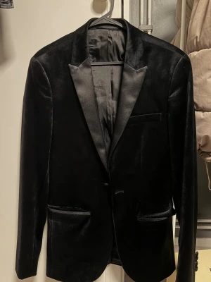 Svart sammetskavaj  - Stilren svart sammetskavaj från Topman med klassisk passform och slimmad siluett. Kavajen har spetsiga slag i satin, tre fickor framtill och en innerficka. Perfekt för dig som vill ha en elegant look med lite extra lyxig känsla.