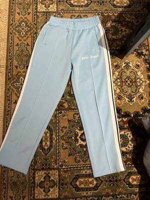 Palm Angels ljusblå mjukisbyxor - Ljusblå mjukisbyxor från Palm Angels med vita sidoränder och resår i midjan. Byxorna har raka ben, fickor på sidorna och logga broderad framtill. . Är storlek M men kan också passa på dom som har S,, kan också tänka mig byte 