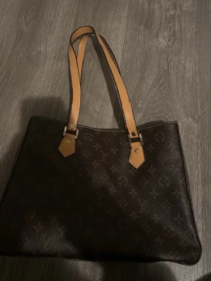 Louis Vuitton Monogram handväska - Säljer en klassisk Louis Vuitton handväska i brunt monogrammönstrat canvas med ljusbruna läderhandtag. Väskan har dragkedja upptill och rymligt innerfack med samma ikoniska mönster. Perfekt för dig som gillar lyxiga accessoarer och tidlös design.