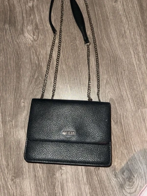 Svart axelväska från Guess - Snygg svart axelväska från Guess med kedjerem och strukturmönstrat konstläder. Väskan har ett stilrent och klassiskt utseende med logotyp i silver framtill. Perfekt storlek för det viktigaste och enkel att matcha med olika outfits.