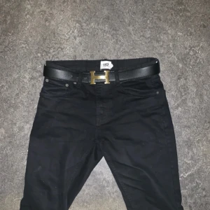 Svart och vitt vändbart bälte med guldfärgat spänne - Snyggt vändbart bälte i svart och vitt med ett stilrent guldfärgat H-spänne. Perfekt för att matcha olika outfits. Tillverkat i skinn för en lyxig känsla.