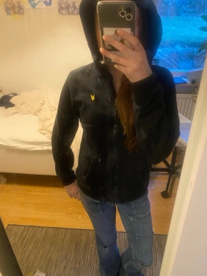 Svart hoodie från Lyle & Scott - Svart hoodie med dragkedja från Lyle & Scott. Klassisk modell med huva och två fickor framtill. Ikonisk gul logga på bröstet. Mjuk och skön bomullsblandning, perfekt för en avslappnad stil.