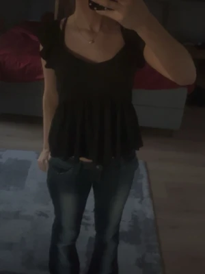 Svart volangtopp med kort ärm - Snygg svart topp med volang nertill och korta ärmar. Toppen har en v-ringad halsringning och är lätt utsvängd för en luftig känsla. Perfekt att styla med jeans eller kjol för en trendig look.