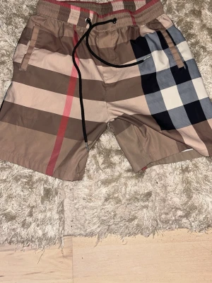 Rutiga badbyxor med dragsko - Snygga badbyxor med klassiskt rutmönster i beige, brunt, svart, vitt och rött. De har elastisk midja med svart dragsko och sidofickor. Perfekta för stranden eller poolen och ger en trendig look.