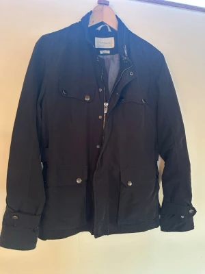 Svart vindjacka från Morris - Stilren svart vindjacka från Zara med fyra fickor framtill, knappar och dragkedja. Jackan har hög krage och justerbara detaljer i midjan samt vid ärmsluten. Perfekt för dig som vill ha en klassisk och funktionell jacka med snygg passform.