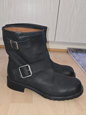 Svarta bikerkängor med spännen - Säljer ett par svarta bikerinspirerade kängor i skinn med två silvriga spännen på sidan. Kängorna har rund tå, grov sula och ett klassiskt, stilrent utseende. Perfekta för dig som vill ha en tidlös look.