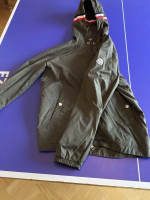 Grön Moncler vindjacka med huva - Stilren grön vindjacka från Moncler med huva och dragkedja. Jackan har Moncler-logga på ärmen, två fickor med tryckknappar och elastiska muddar. Huvan har snygga ränder i rött och vitt. Perfekt för dig som vill ha en lätt och sportig jacka. Bara än änd ett par få tal gånger