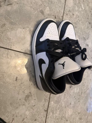 Svarta och vita Air Jordan 1 Low - Säljer ett par stilrena Air Jordan 1 Low i svart och vitt läder. Skorna har klassisk låg siluett, svart swoosh på sidan och ikoniska Jumpman-loggan på plösen. Perfekta för dig som vill ha en clean och tidlös sneaker med streetkänsla. 