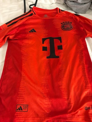Röd FC Bayern München matchtröja - Snygg röd FC Bayern München matchtröja från Adidas med svarta detaljer och klubbmärke på bröstet. Tröjan har klassiska Adidas-ränder på axlarna och stor svart logga på framsidan. Tillverkad i lätt och ventilerande funktionsmaterial, perfekt för fotbollsträning.