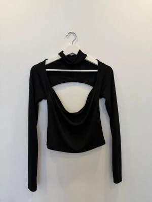 Svart långärmad topp Gina Tricot - Trendig svart långärmad topp från Gina Tricot med öppen rygg och snygg draperad urringning. Toppen har en croppad passform och är tillverkad i ett stretchigt syntetmaterial som sitter bekvämt. Perfekt för dig som vill sticka ut med en unik detalj.