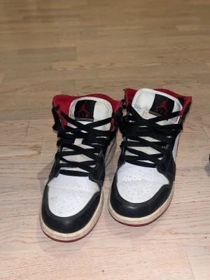 Air Jordan 1 Mid svart/vit/röd - Säljer ett par Air Jordan 1 Mid sneakers från Nike i svart, vitt och rött. Skorna har klassisk hög siluett, svart swoosh, röda detaljer och vit tåbox. Ovandelen är i läder och de har svarta skosnören samt röd insida. Perfekta för dig som gillar streetwear.