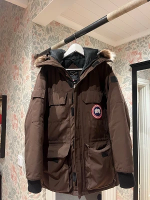 Brun parkas från Canada Goose - Säljer en brun parkas från Canada Goose med ikonisk logga på bröstet, flera stora fickor och svart innerfoder. Jackan har huva med pälskant och muddar vid ärmsluten. Perfekt för kalla dagar och riktigt snygg streetstil. Det enda felet är på den sista bilden!