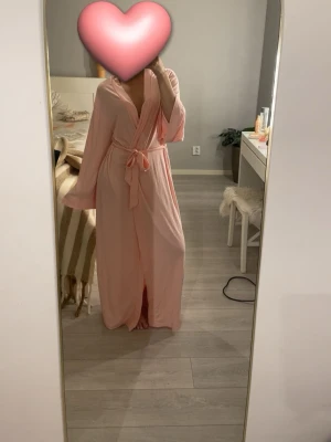 Ljusrosa lång morgonrock med knytskärp - Mysig och stilren ljusrosa morgonrock i lång modell med vida ärmar och knytskärp i midjan. Perfekt för att mysa hemma. Tunt och mjukt material som ger en avslappnad känsla. Liknande Alobha morgonrocken! 