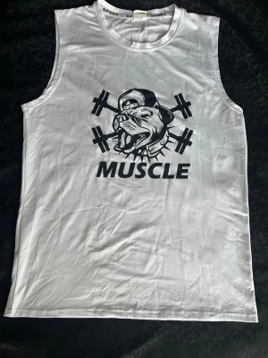 Vit linnetopp med tryck MUSCLE L - Vit ärmlös linnetopp med stort svart tryck av en hund med keps och vikter samt texten MUSCLE på bröstet. Perfekt för dig som gillar gym eller träning. Materialet känns som bomull eller polyester och toppen har en normal passform.