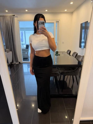 Svart långkjol med slits - Stilren svart långkjol med hög midja och slits på sidan. Kjolen har bälteshällor och en figurnära passform som framhäver siluetten. Perfekt att matcha med en croppad topp för en trendig look.