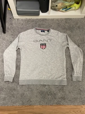 Grå sweatshirt från GANT - Snygg grå sweatshirt från GANT med broderad logga och sköld på bröstet. Tröjan har rund halsringning, ribbade muddar och klassisk passform. Perfekt för en avslappnad och stilren look. Pris kan sänkas 