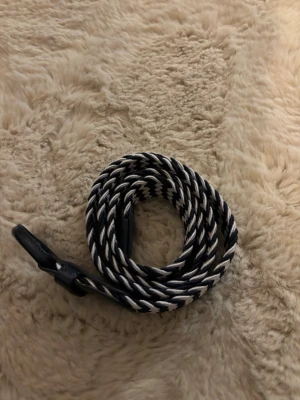 Svartvitt flätat bälte - Snyggt flätat bälte i svart och vitt med smal design. Bältet har ett randigt mönster och är perfekt för att ge din outfit en cool touch. Spännet och ändarna är i svart material.