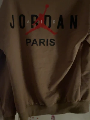 Brun Jordan Paris sweatshirt - Säljer en brun sweatshirt från Jordan Paris-kollektionen. Tröjan har rund hals, långa ärmar och tryck med svart text och röd Jumpman-logga på bröstet. Perfekt för dig som gillar streetwear och sportig stil.